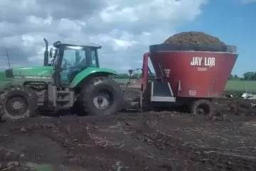 Tractor verde remolcando mezcladora de alimento Jay-Lor roja cargada con forraje, trabajando en terreno fangoso bajo cielo nublado en ambiente rural