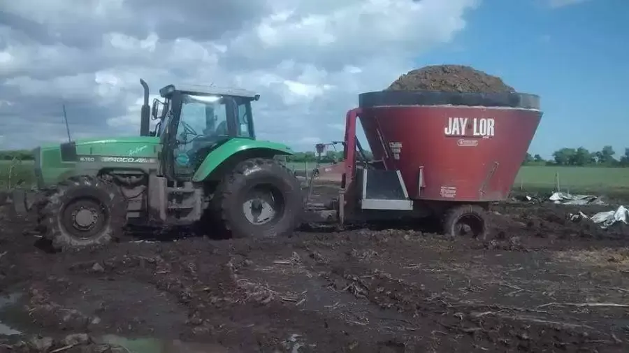 Tractor verde remolcando mezcladora de alimento Jay-Lor roja cargada con forraje, trabajando en terreno fangoso bajo cielo nublado en ambiente rural