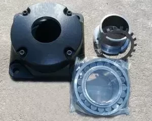 Componentes mecánicos metálicos incluyendo carcasa cuadrada con orificio central, adaptador cilíndrico y rodamiento sobre superficie de concreto