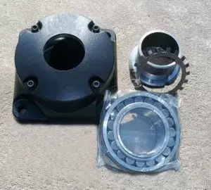 Componentes mecánicos metálicos incluyendo carcasa cuadrada con orificio central, adaptador cilíndrico y rodamiento sobre superficie de concreto