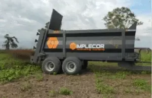Remolque agrícola marca Implecor de color negro con doble eje de ruedas y caja basculante elevada, ubicado en campo rural con árboles de fondo